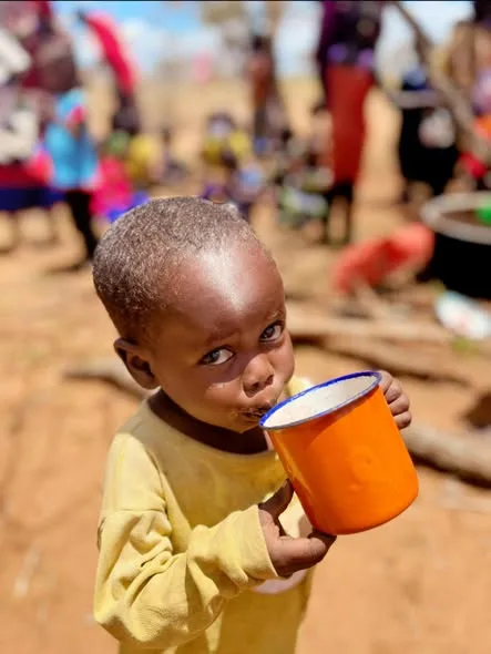 Litet barn dricker ur en orange mugg vid en matutdelning i en by i Kenya